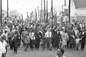 Dr. Martin Luther King, fourth from right, waves as an Army observation plane flies over flag-carrying civil rights demonstrators near Montgomery, Ala., March 24, 1965.  To the right of King is his wife Coretta Scott King, and behind his upraised arm is singer Harry Belefonte. This is the fourth day of the Selma to Montgomery civil rights march.  (AP Photo)